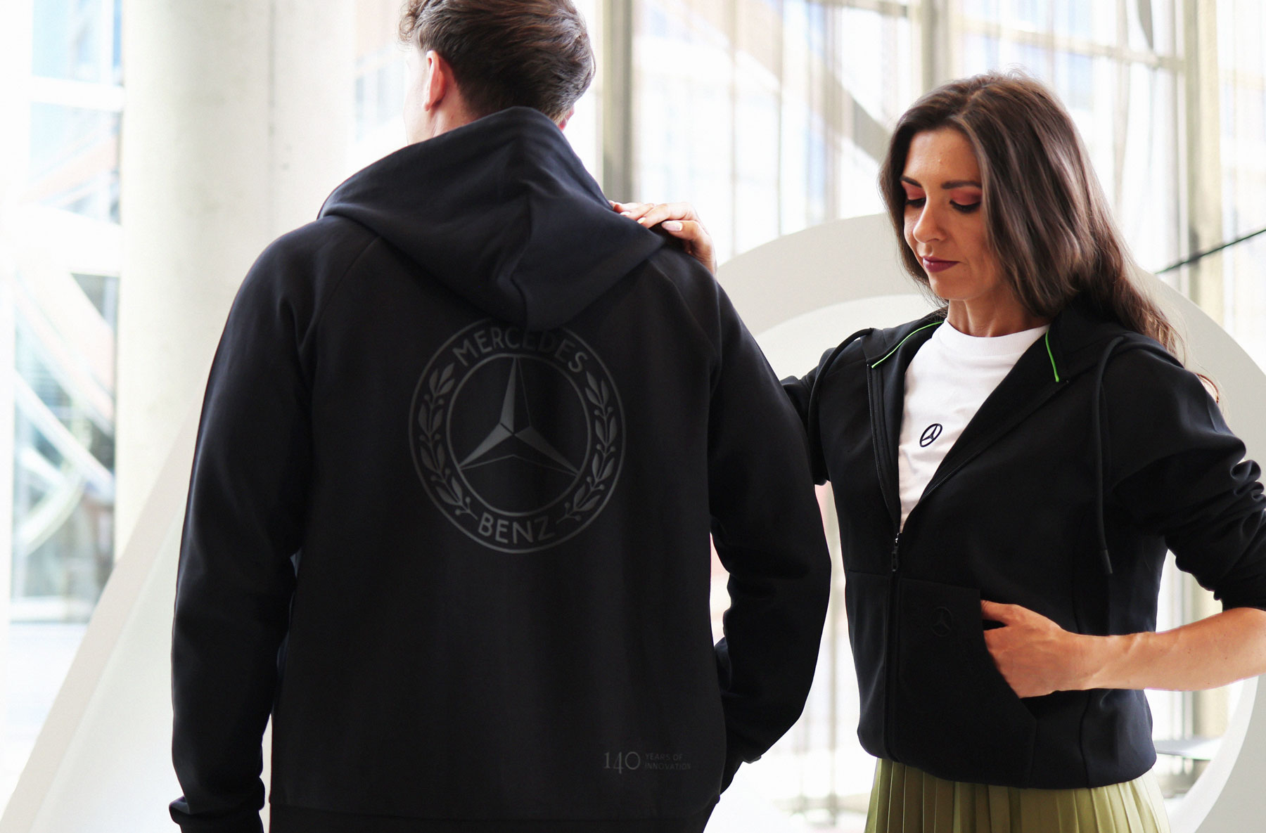 Mercedes-Benz 140 Years Collection Schwarze Sweatshirt-Jacke der Mercedes-Benz „140 Years of Innovation Collection“. Das Kleidungsstück wird von zwei Personen präsentiert, die Rückseite zeigt ein großes Mercedes-Benz Logo.
