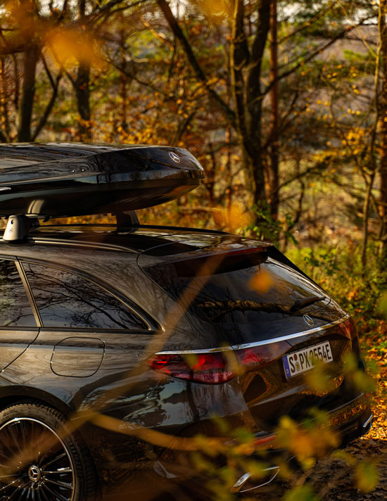 Abgebildet wird ein schwarzer Mercedes-Benz mit montierter Dachbox vor einem herbstlichen Waldhintergrund.